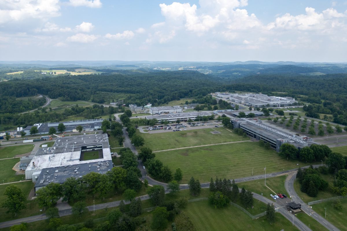 Aerial view of a large industrial or office complex surrounded by green fields, trees, and roads, with a forested landscape and hills in the background under a partly cloudy sky.