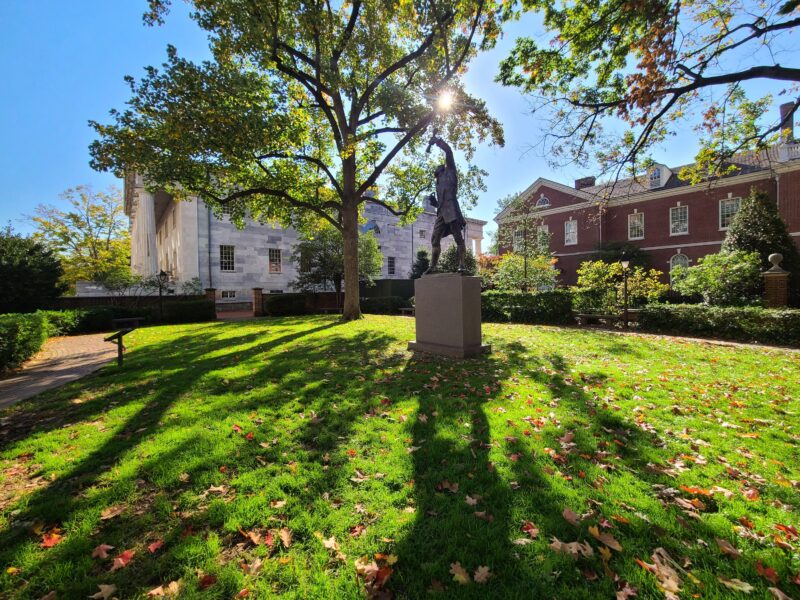 A statue stands on a pedestal in a grassy courtyard, surrounded by trees, historic brick and stone buildings, and scattered autumn leaves.