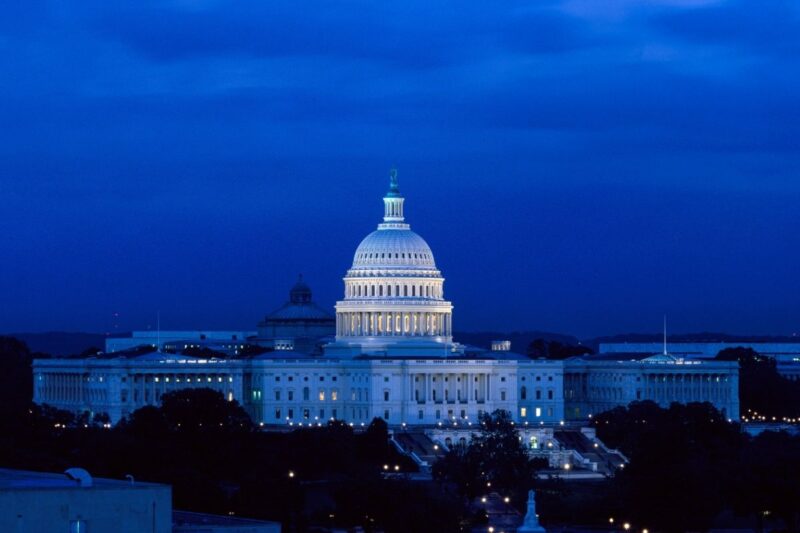 The United States Capitol building is illuminated at night against a dark blue sky, with trees and other buildings in silhouette in the foreground.