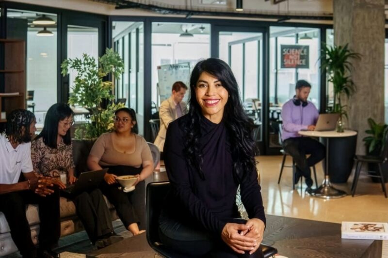 Woman in black top sits and smiles at the camera in a modern office space with other people working and talking in the background.