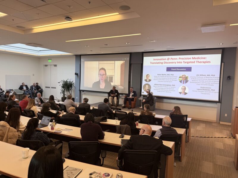 Audience watches a panel discussion on "Innovation @ Penn: Precision Medicine" with speakers seated on stage and a large screen displaying their names and photos.