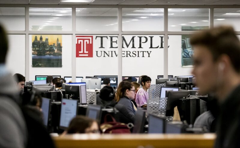 Students work at computer stations in a large lab at Temple University, with the university logo visible on the wall in the background.