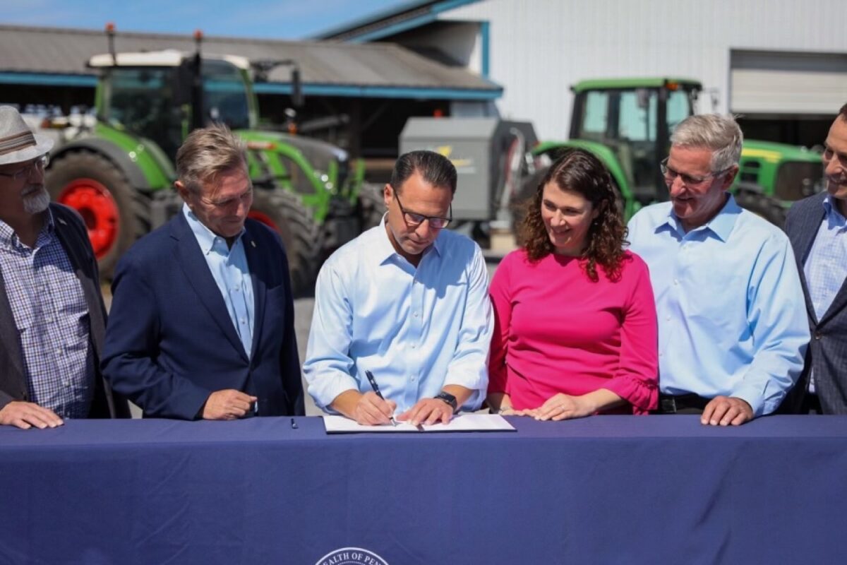 A group of five adults stand behind a table as a man in the center signs a document; tractors and a building are visible in the background.