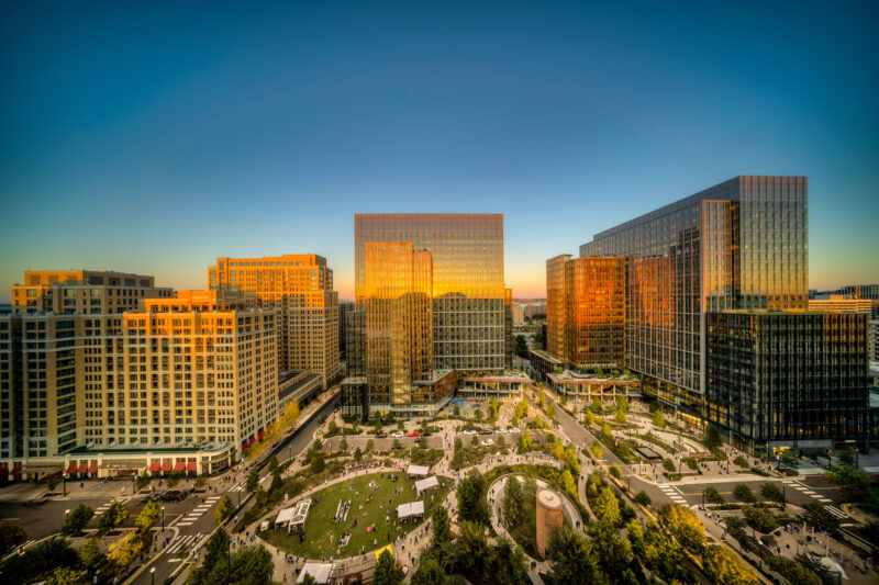 Aerial view of modern office buildings with glass facades reflecting sunset, overlooking a city park with people and pathways below.