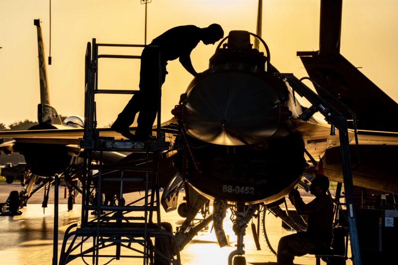 Two people perform maintenance on a fighter jet at sunset, with one standing on a platform and the other working below the aircraft in a hangar.