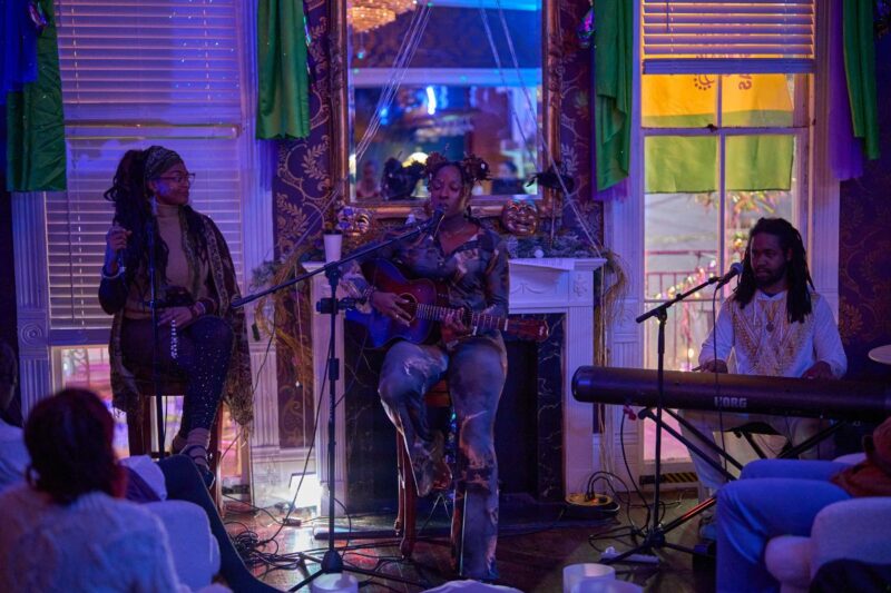 Three musicians perform indoors: one sings and plays guitar, one plays keyboard, and one holds a microphone. The room has colorful lighting, curtains, and seated audience members.
