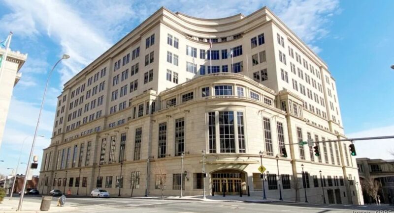 A large, multi-story beige office building with many windows sits on a street corner under a partly cloudy sky.