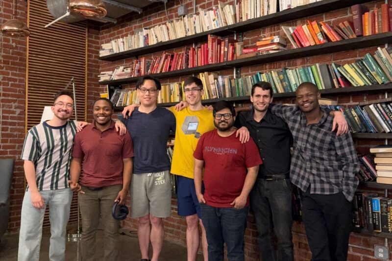 Seven people stand side by side in front of a wall of bookshelves, smiling at the camera in a casual indoor setting.