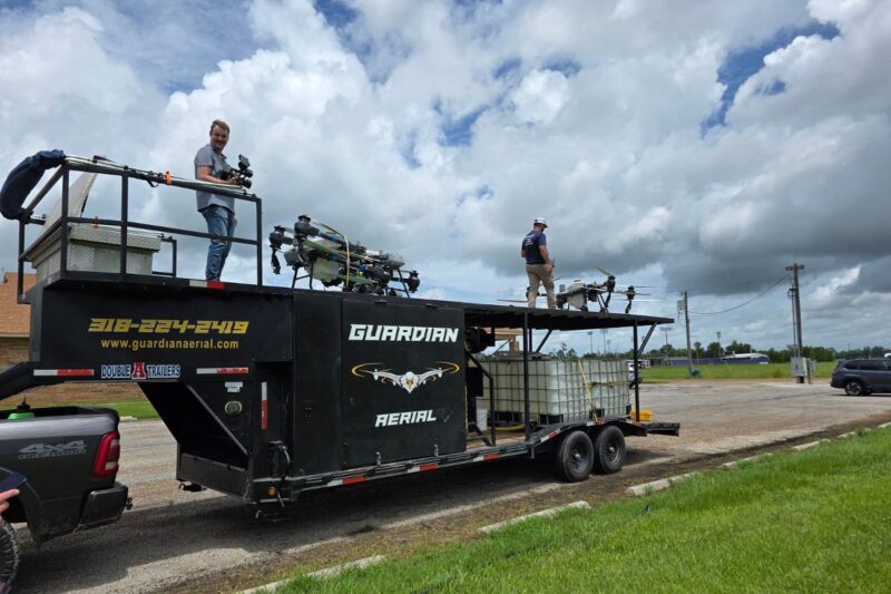 Two people stand on a large trailer labeled "Guardian Aerial" with drone equipment, parked on a grassy lot under a partly cloudy sky.