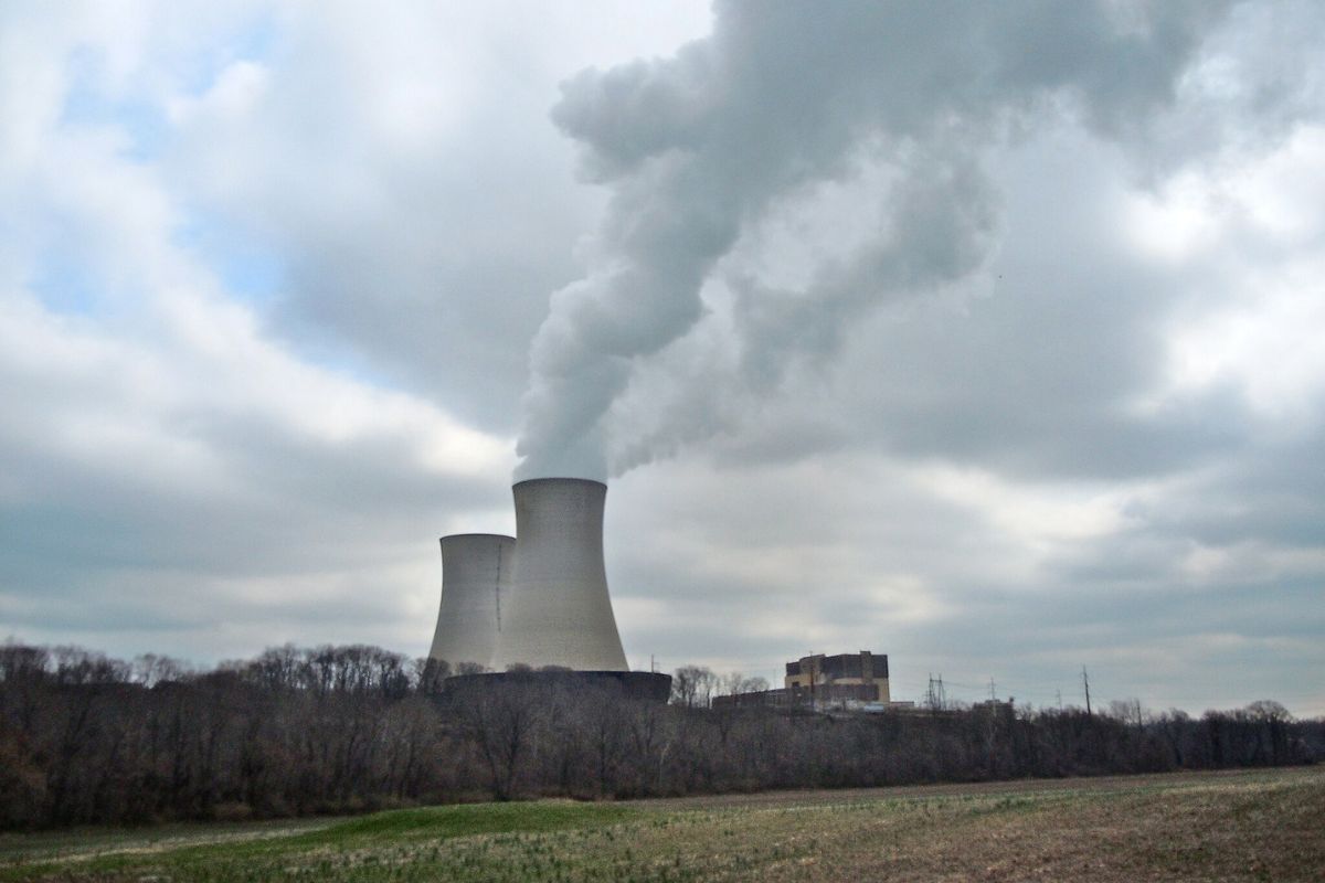 Two large cooling towers emit steam at a power plant next to a wooded area and open field under a cloudy sky.