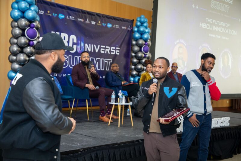 A Black man in a jacket with a V on it holds a microphone and a plaque in front of a stage