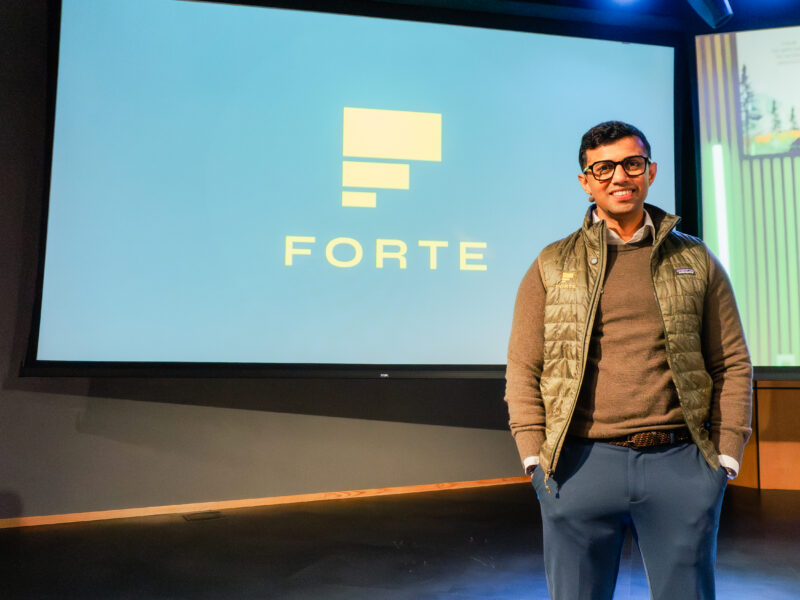 A South Asian man stands in front of a blue screen that says Forte