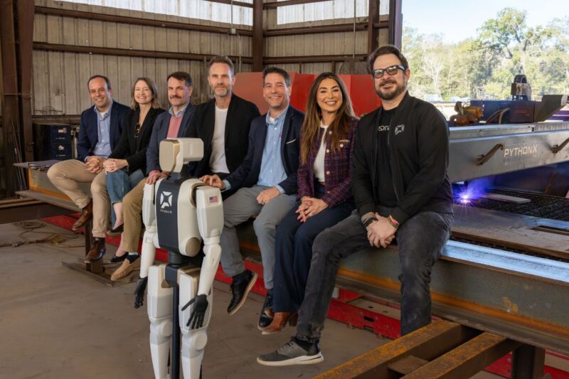 A group of seven people sits on metal beams inside an industrial facility, posing with a white humanoid robot in front of them.