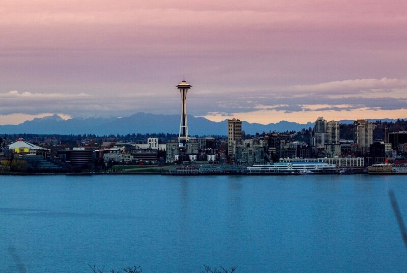 A city skyline with a prominent observation tower beside a large body of water, with mountains visible in the background under a cloudy sky.
