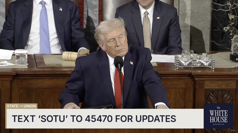 A man in a suit stands at a podium, speaking into microphones during a formal event, possibly addressing topics like Trump’s ‘ratepayer protection pledges’ for data centers. Text reads: “TEXT 'SOTU' TO 45470 FOR UPDATES.”.
