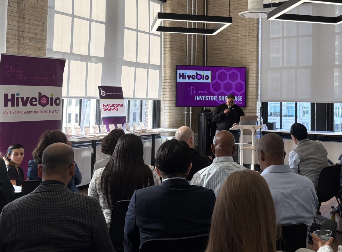 People attend a HiveBio investor showcase event in a modern, well-lit room with large windows and presentation screens displaying event branding.