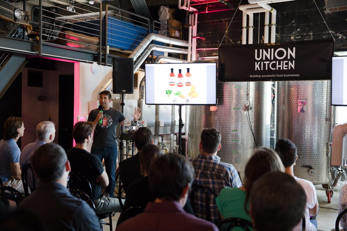 A speaker gives a presentation to an audience at Union Kitchen, with product images displayed on screens and industrial equipment in the background.