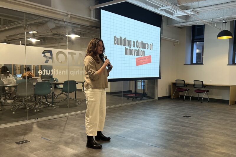 A woman holding a microphone stands in front of a screen displaying the words "Building a Culture of Innovation" in a modern office or conference space.