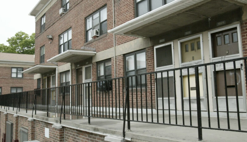The exterior of a public housing complex. A railing is in the foreground with entrance doors in the back