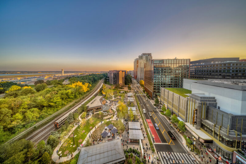 Aerial view of a cityscape at sunset with office buildings on the right, a park and train tracks on the left, and a road with cars and people in the foreground.