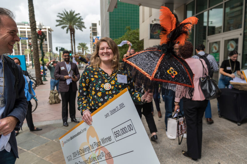 A woman smiles while holding a large check for $50,000 and a decorative umbrella at an outdoor event with people in the background.