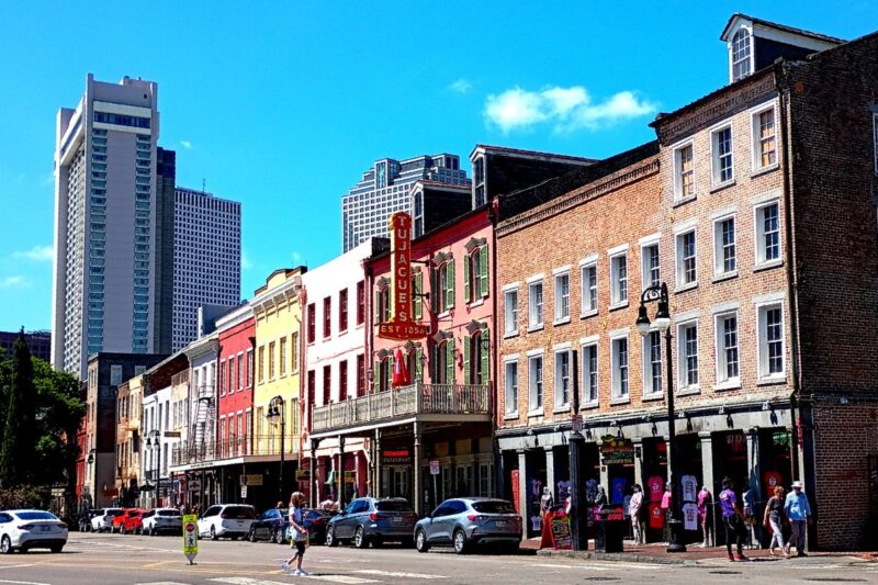 A row of historic brick buildings and shops lines a busy street in New Orleans, with modern skyscrapers visible in the background under a clear blue sky.