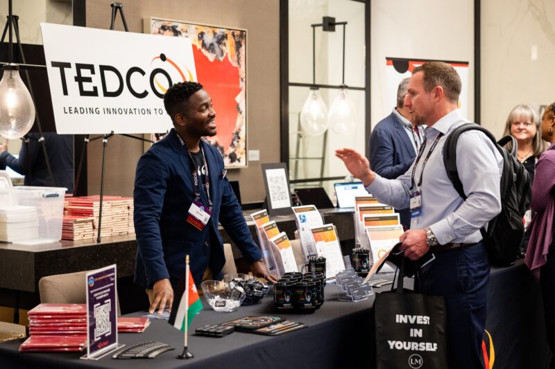 Two men converse at a TEDCO booth during a conference; branded materials and informational brochures are displayed on the table.