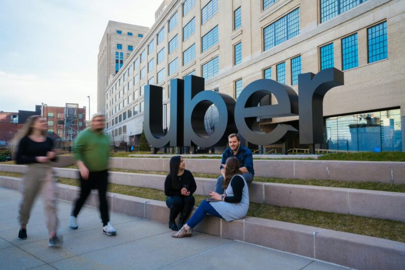 Four people sit and talk near a large "Uber" sign outside an office building, while two others walk past in the foreground.