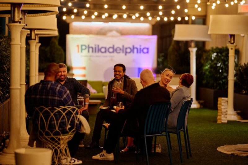 A group of people sit and talk at an outdoor event space under string lights, with a "1Philadelphia" banner in the background.