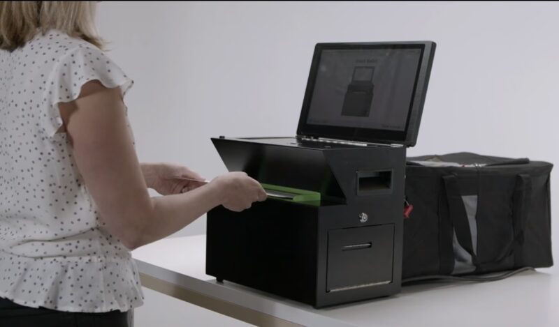 A person inserts a stack of papers into a black ballot scanning machine with an attached laptop screen displaying instructions.