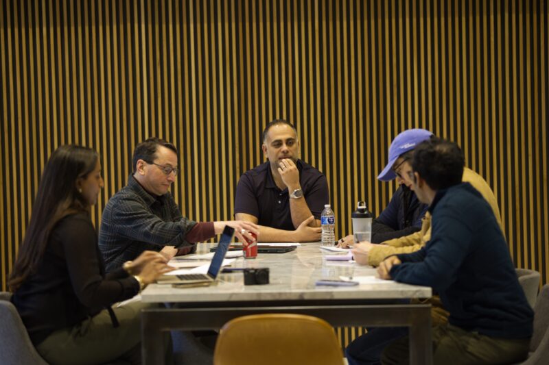 Five people sit around a table having a discussion, with notebooks, laptops, and drinks in front of them against a backdrop of vertical wooden slats.