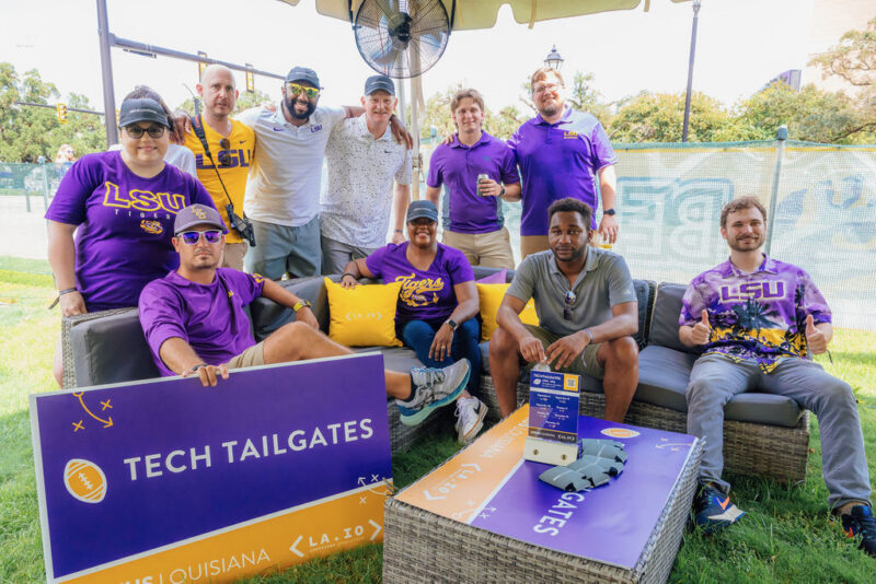 A group of people wearing LSU apparel pose together outdoors at a tech tailgates event, seated on couches with event signage and a fan in the background.