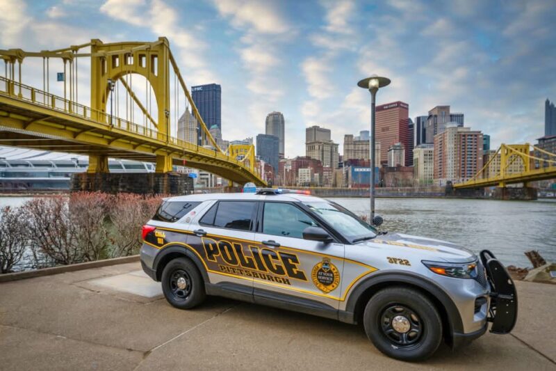 A Pittsburgh Police SUV is parked by the river with a yellow bridge and city skyline in the background under a cloudy sky.