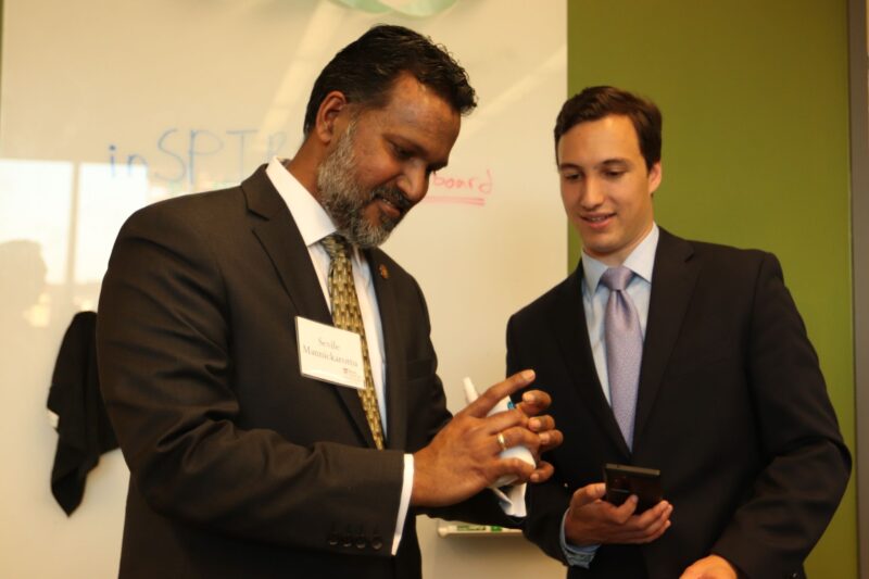 Two men in business suits stand in front of a whiteboard, one holding a marker and writing on a notepad while the other looks on holding a phone.