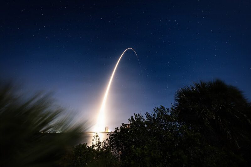 A rocket launches into the night sky, leaving a bright arc of light above silhouetted trees and water under a starry sky.