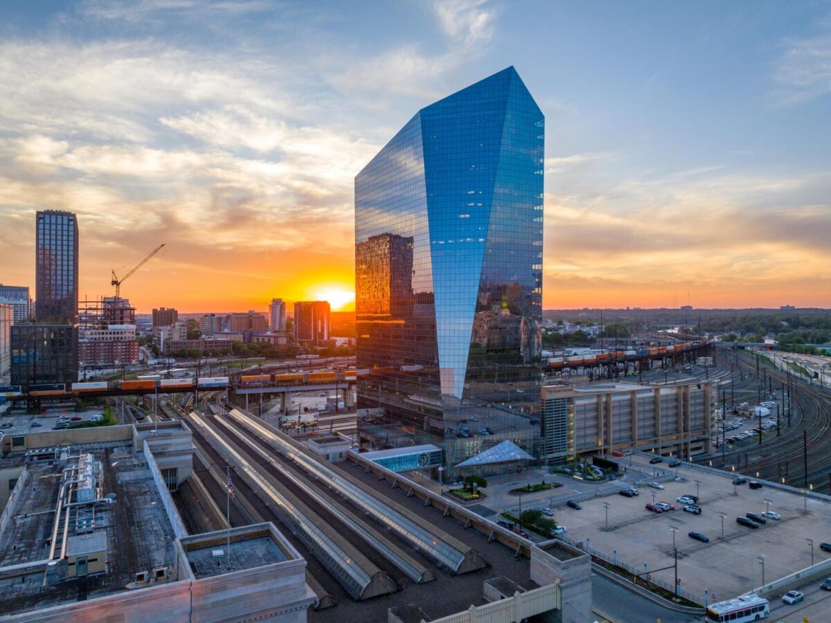 A modern glass skyscraper reflects the sunset beside a rail station with multiple train tracks, surrounded by city buildings and construction cranes.