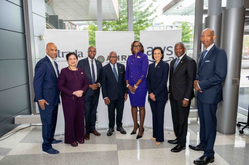 Eight professionally dressed individuals stand in a row posing for a group photo in front of branded banners inside a modern building.