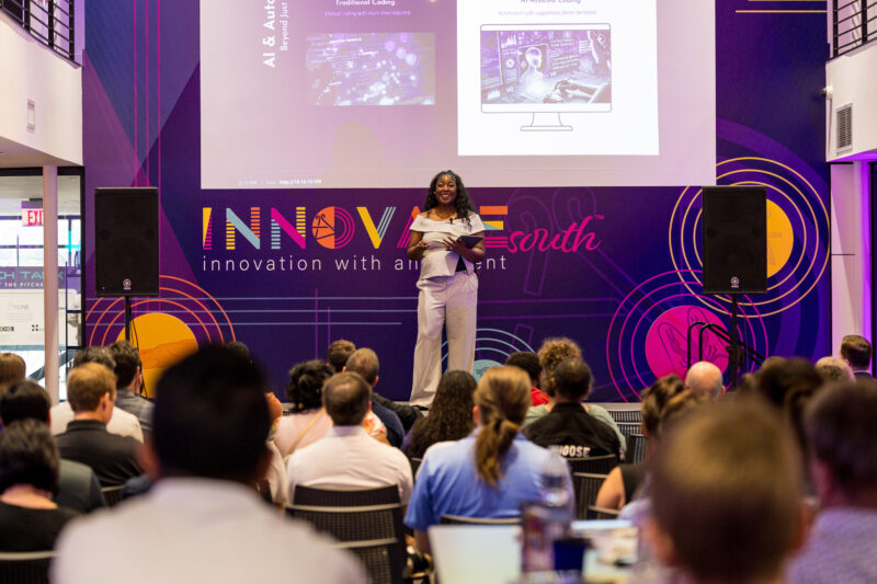 A woman stands on stage speaking to an audience at the Innovate South event, with AI-related slides displayed behind her.