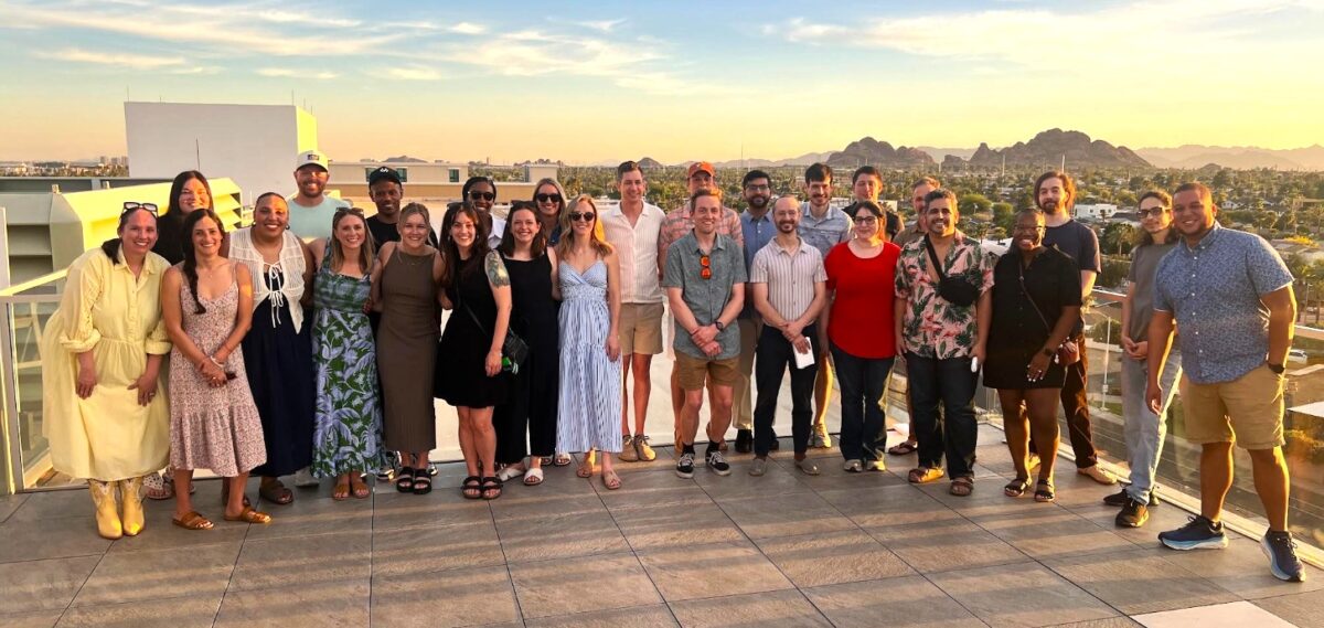 A group of about 25 people stand together on a rooftop at sunset, with mountains and cityscape in the background.