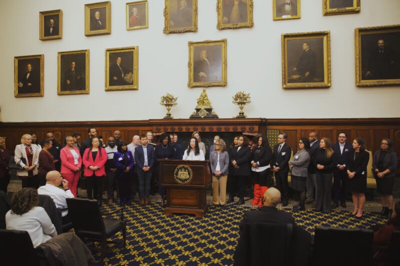A group of people stands behind a podium in a formal room with framed portraits on the wall and patterned carpet, while an audience listens.