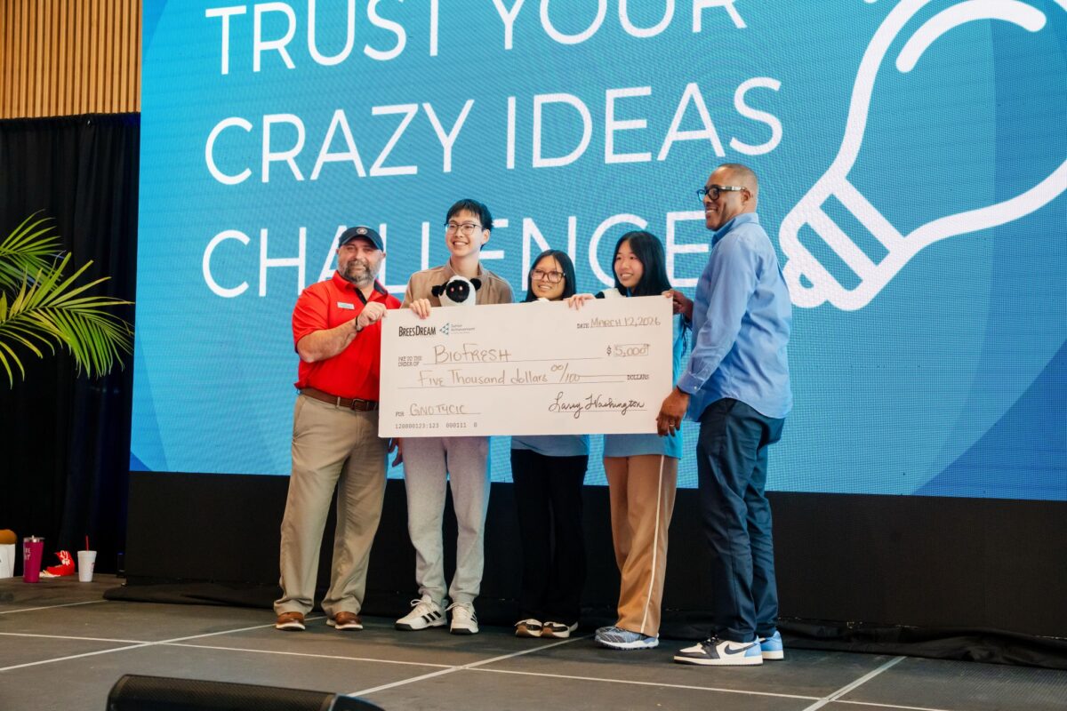 Five people stand on stage holding a large check for $5,000, with a blue background reading "Trust Your Crazy Ideas Challenge.