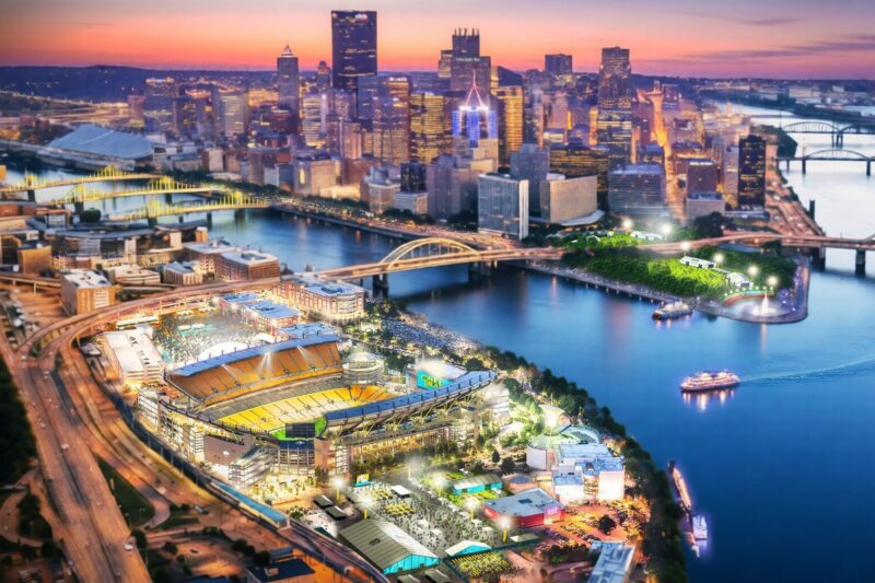 Aerial view of a brightly lit stadium by a river at sunset, with a city skyline and multiple bridges in the background.