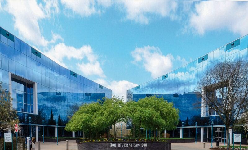 Modern office buildings with reflective glass facades surround a courtyard with green trees; the address "300 RIVERVIEW" is visible on a central sign.