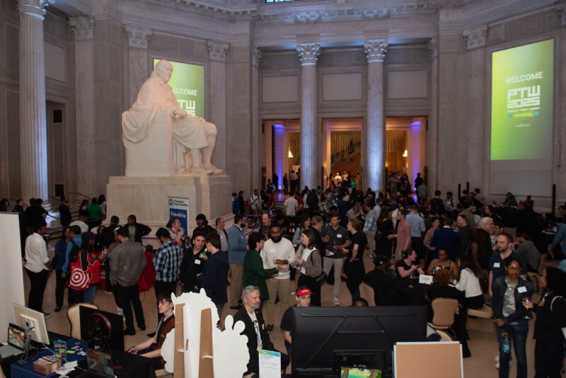 Large crowd gathers at an indoor event in a grand hall with tall columns, a large statue, and projection screens displaying "WELCOME PTW 2025" on the walls.