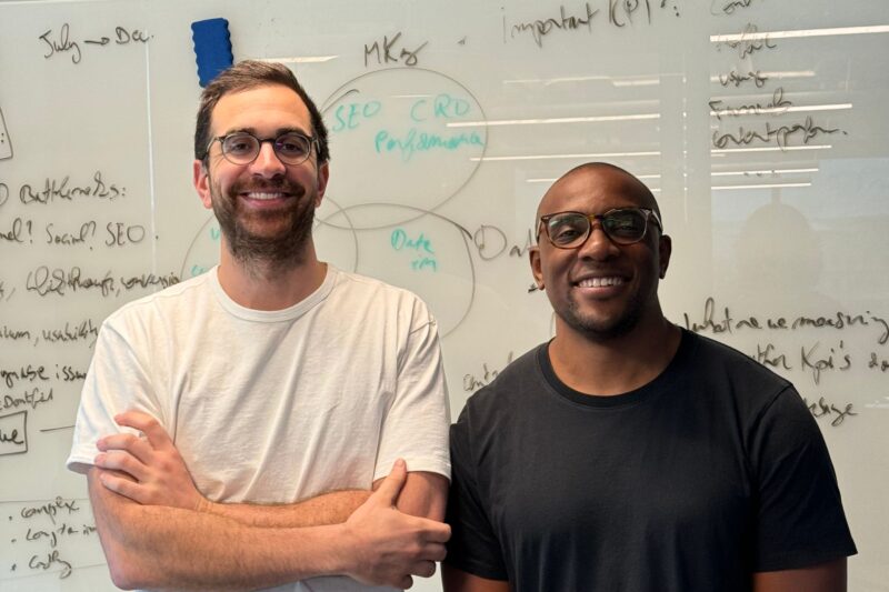 Two people stand smiling in front of a whiteboard covered with handwritten notes and diagrams.