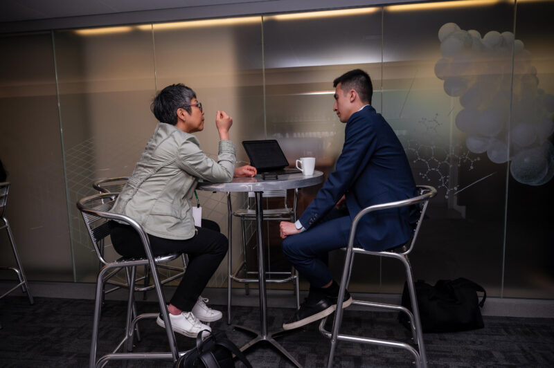 Two people sit at a round table having a conversation, with a laptop and cup on the table in a modern office setting.