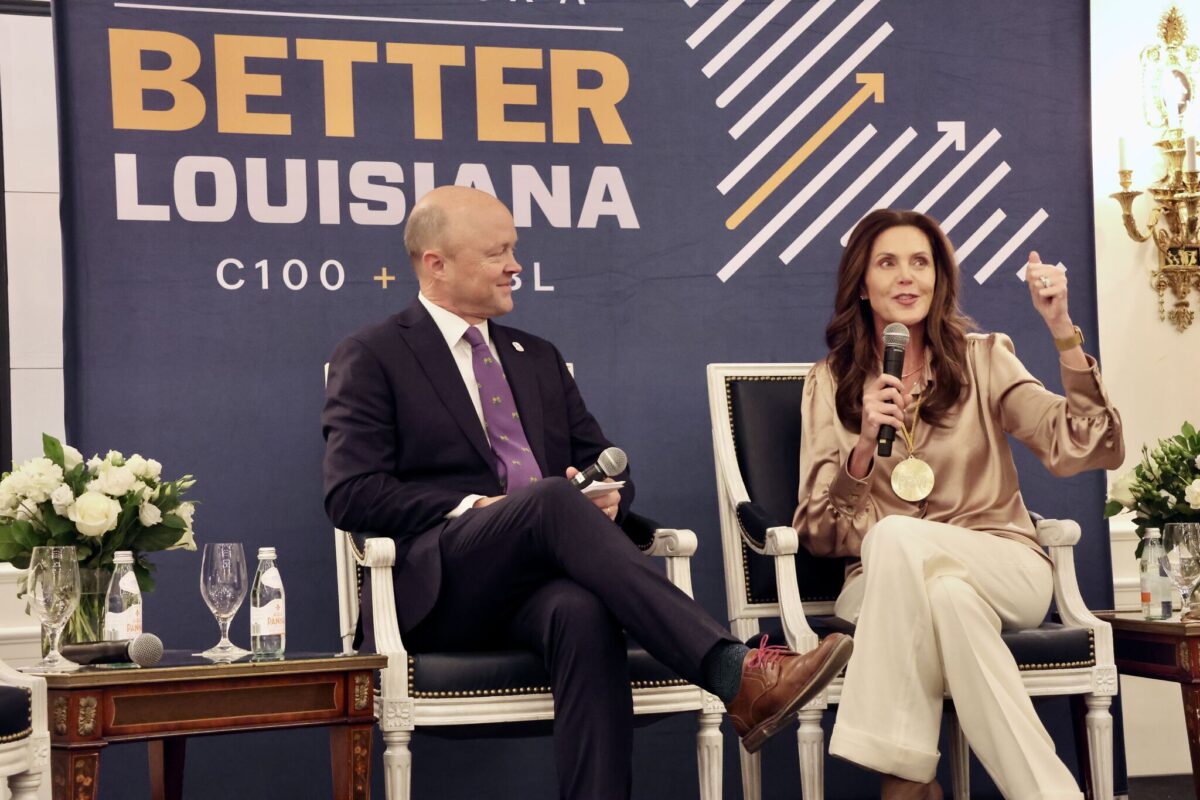 A man and a woman speak on stage at a panel event in front of a "Better Louisiana" banner. The woman is gesturing with her hand while talking.
