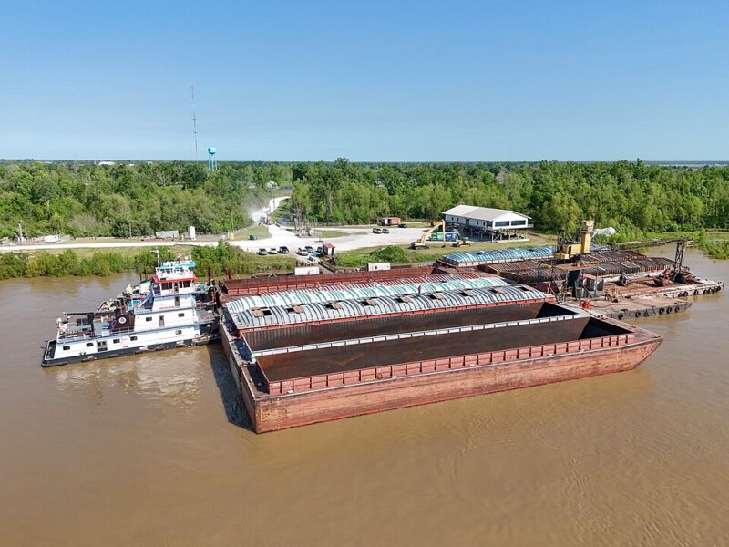 A tugboat pushes a large, empty barge on a brown river near a dock with buildings and trees in the background under a clear blue sky.
