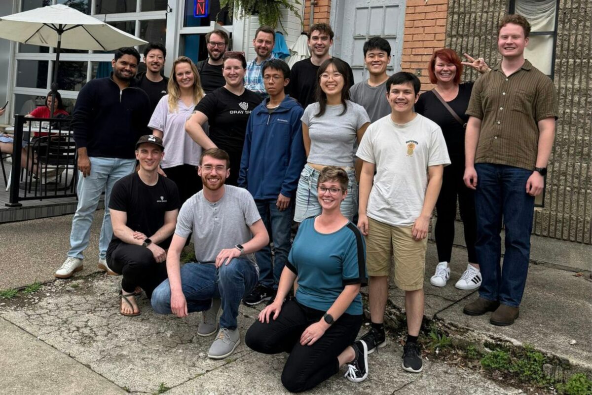 A group of sixteen people pose and smile for a photo outside a building with outdoor seating and umbrellas in the background.