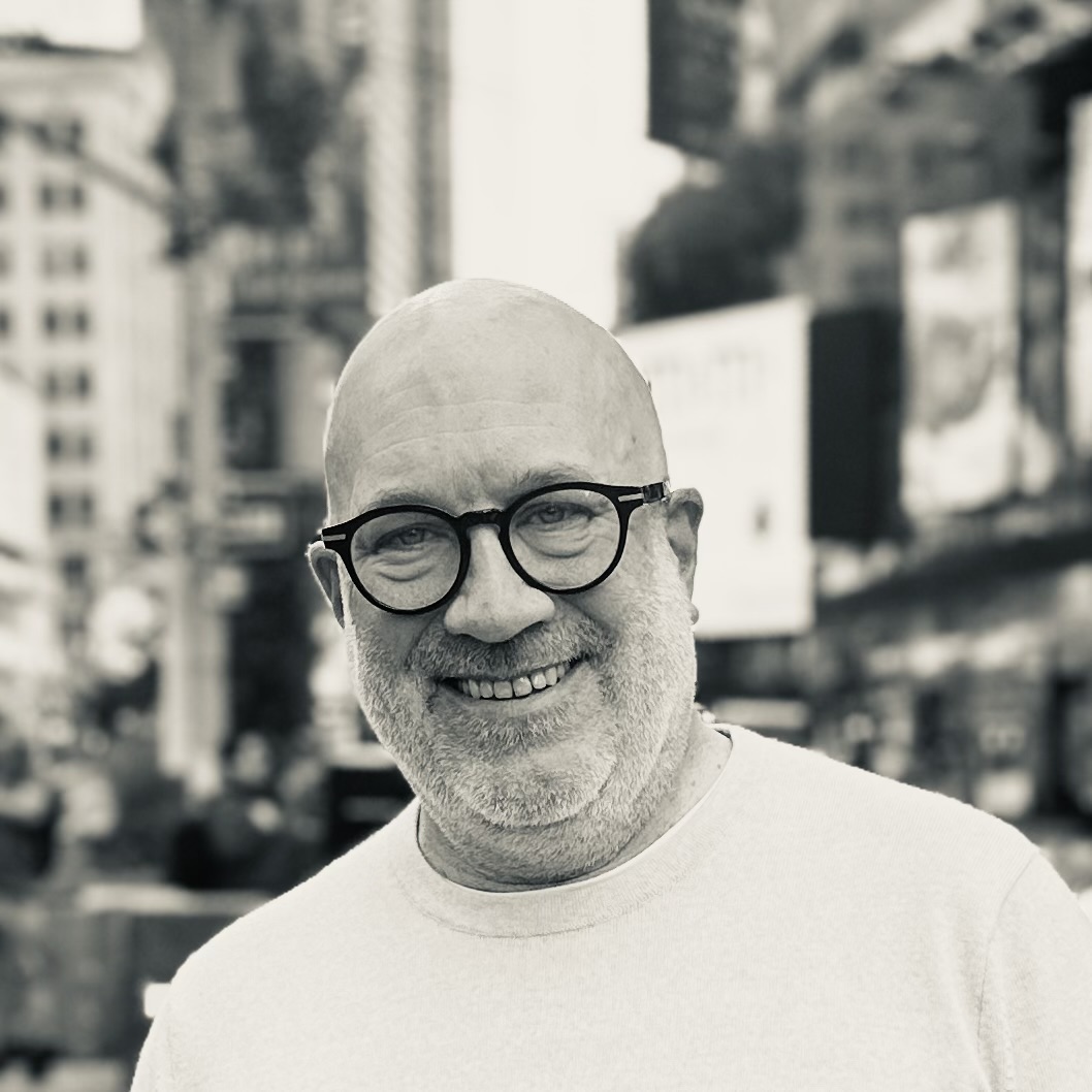 A smiling man with glasses and a beard stands outdoors in an urban setting. The photo is in black and white.